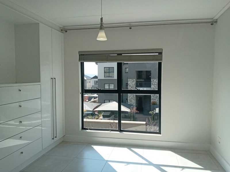 A bright, empty bedroom featuring modern white built-in cupboards, tiled flooring, and a large window with a roller blind.