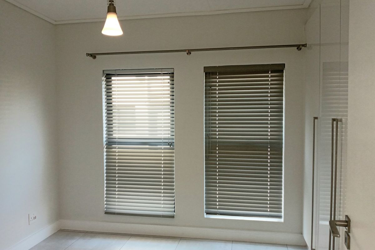 A bright, empty bedroom featuring tiled flooring, built-in cupboards, and dual windows fitted with venetian blinds.