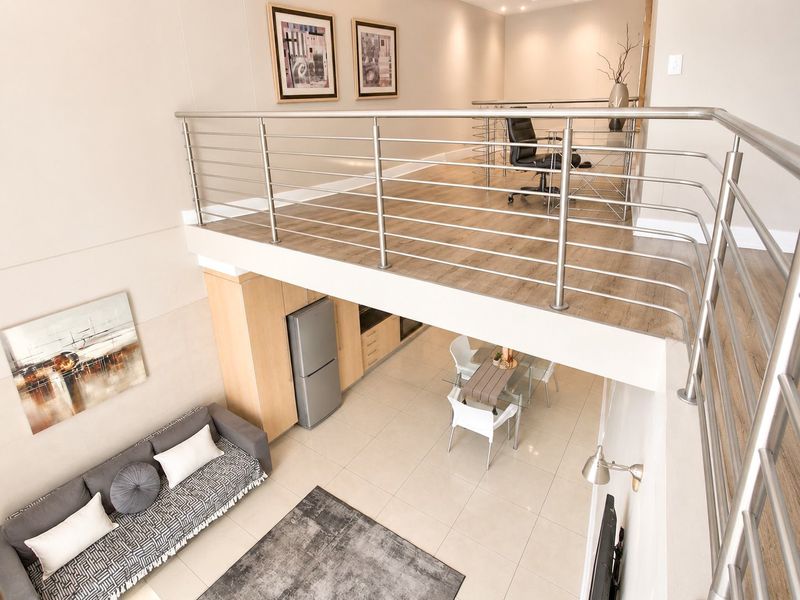 A modern open-plan apartment interior featuring a mezzanine level with a stainless steel balustrade, overlooking a tiled living and dining area.