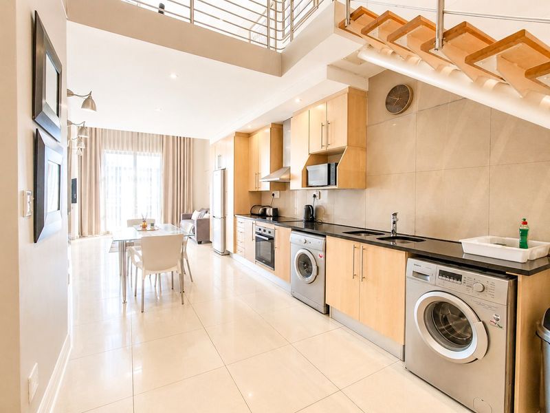 Modern open-plan kitchen and living area featuring light wood cabinetry, stone countertops, integrated appliances, and a staircase leading to an upper level.