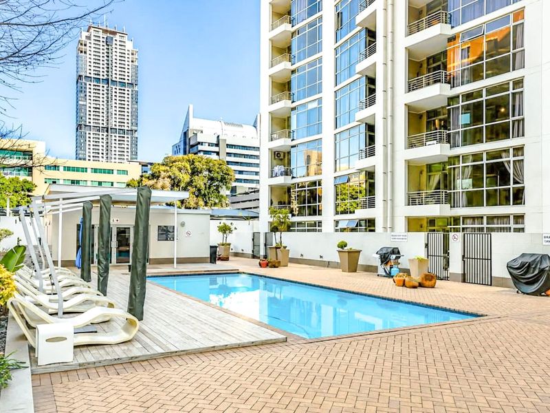 A well-maintained communal swimming pool area featuring timber decking, paved surrounds, and modern apartment building architecture in an urban setting.