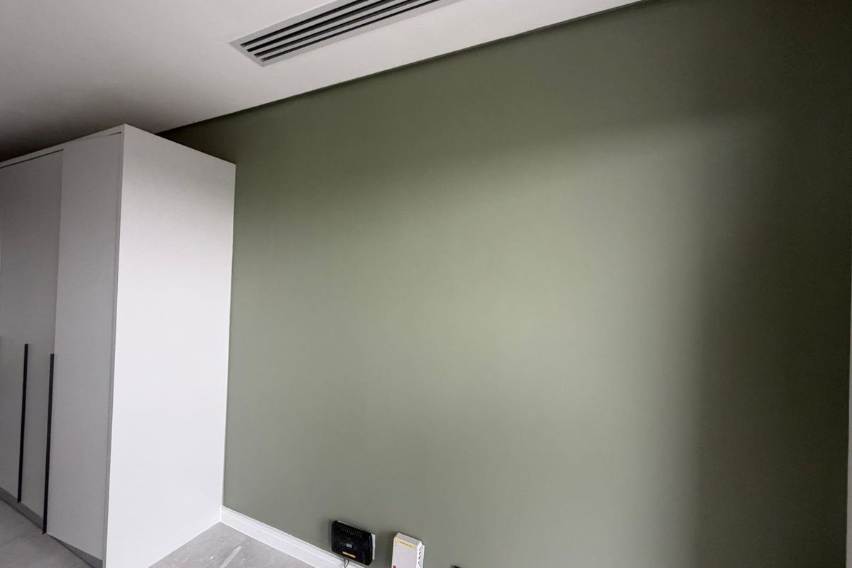 A clean, modern bedroom featuring a built-in wardrobe, neutral green accent wall, and integrated ducted air conditioning.