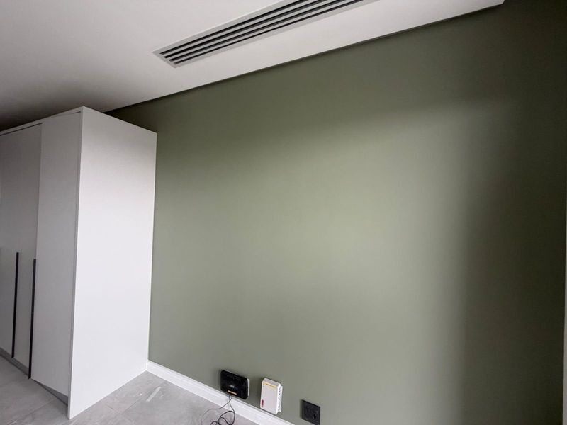 A clean, modern bedroom featuring a built-in wardrobe, neutral green accent wall, and integrated ducted air conditioning.