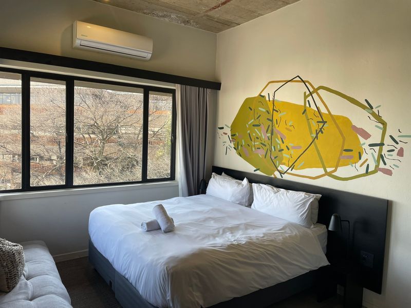 A modern bedroom featuring an exposed concrete ceiling, split-system air conditioning, and a large window with black frames, offering a contemporary urban aesthetic.