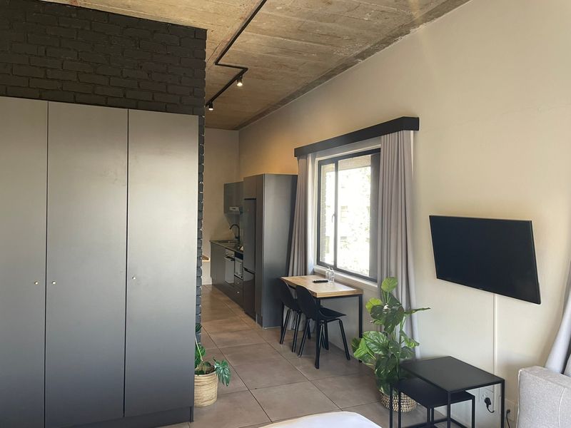Modern open-plan apartment featuring an exposed concrete ceiling, industrial-style track lighting, sleek built-in cabinetry, and a striking black exposed brick feature wall.
