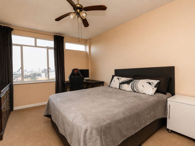 A well-lit bedroom featuring carpeted flooring, a ceiling fan, and a large window offering an elevated city view.
