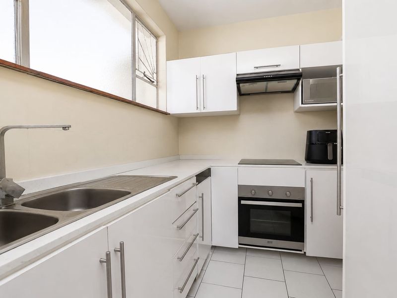 A clean, modern kitchen featuring white cabinetry, stone countertops, stainless steel sink, and integrated appliances including an oven, hob, and extractor fan.
