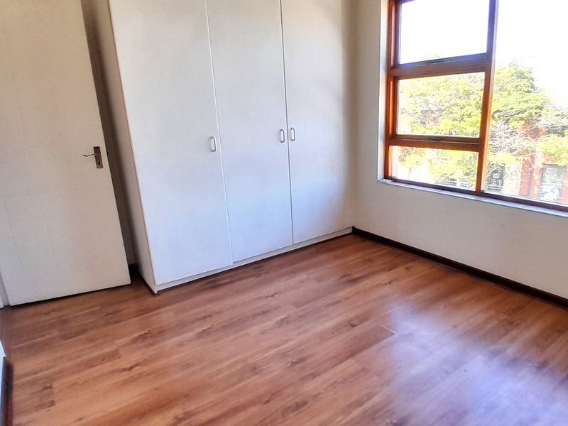 A bright, empty bedroom featuring warm-toned laminate flooring, built-in white cupboards, and a large timber-framed window providing natural light.