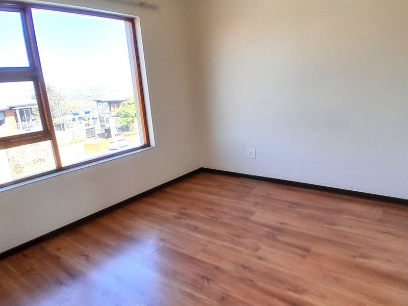A bright, empty bedroom featuring warm-toned laminate flooring, neutral walls, and a large aluminium-framed window providing natural light.