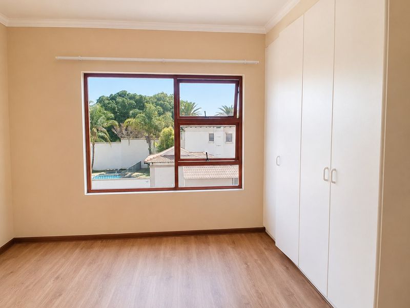 A bright, empty bedroom featuring modern laminate flooring, neutral wall tones, and ample built-in cupboard storage with a view of the surrounding estate.