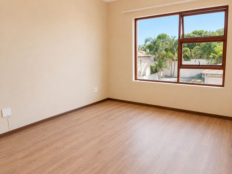 A bright, empty bedroom featuring light-toned laminate flooring, neutral walls, and a large window with a pleasant garden view.