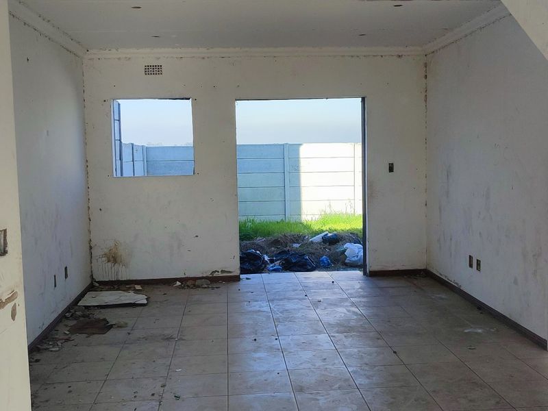 An unfinished open-plan living area featuring tiled flooring, a staircase, and direct access to the rear yard, requiring completion and cosmetic finishing.