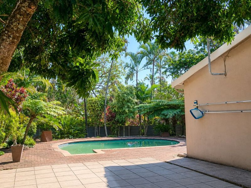 A private outdoor pool area featuring a paved patio, lush tropical landscaping, and a boundary wall, offering a serene backyard retreat.