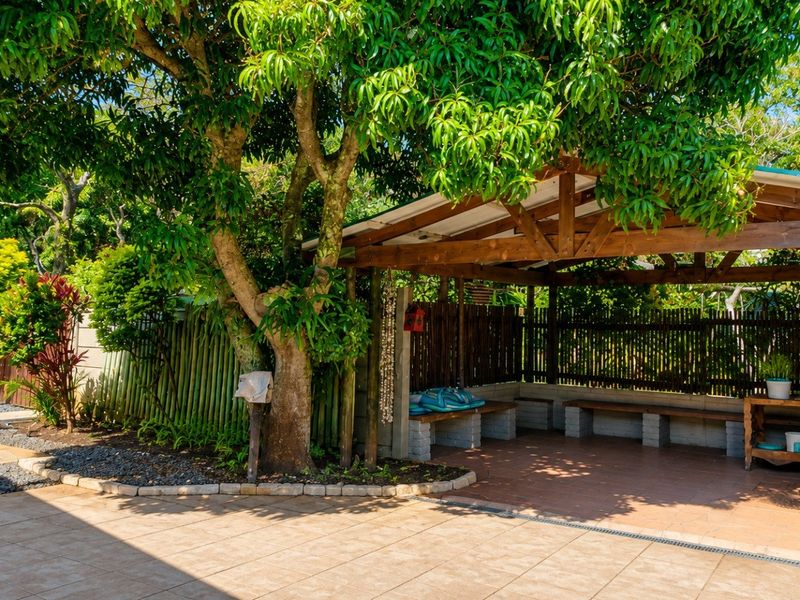 A covered outdoor patio area featuring a timber pergola, built-in masonry bench seating, and tiled flooring, set within a landscaped garden environment.