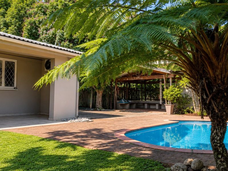 A well-maintained outdoor area featuring a swimming pool, brick-paved patio, and a covered seating area surrounded by established garden greenery.