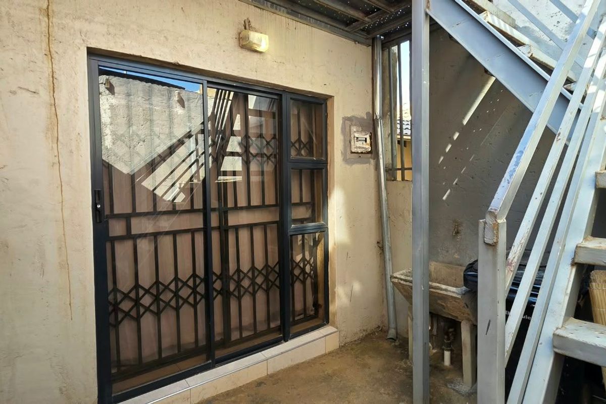 Rear exterior area featuring a sliding door with security gate, metal staircase, and a concrete utility sink, showing signs of wear and weathering.