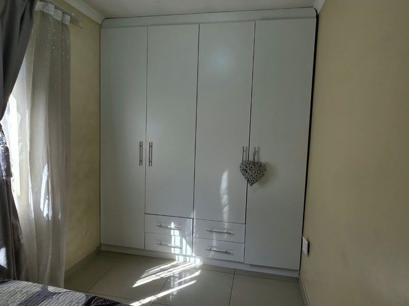 A bedroom featuring a modern white built-in wardrobe with integrated drawers and tiled flooring, illuminated by natural light from the adjacent window.