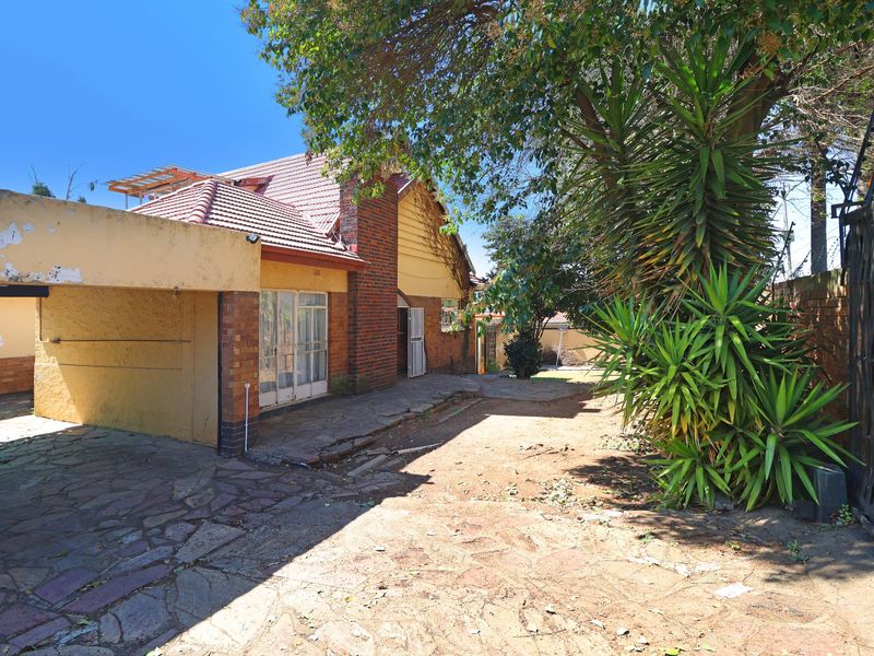 Rear exterior view of the property featuring a paved patio area, attached carport, brick chimney, and established garden with security fencing.