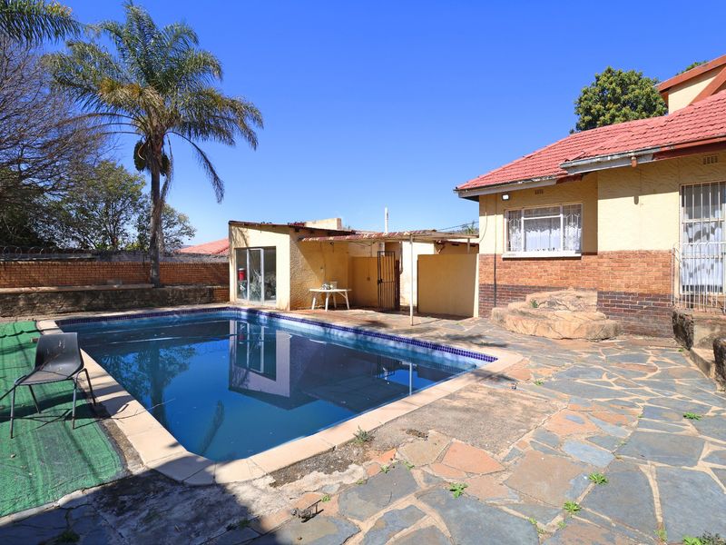 Rear exterior view featuring a swimming pool, paved patio area, and brick boundary wall, showing signs of wear and weathering.
