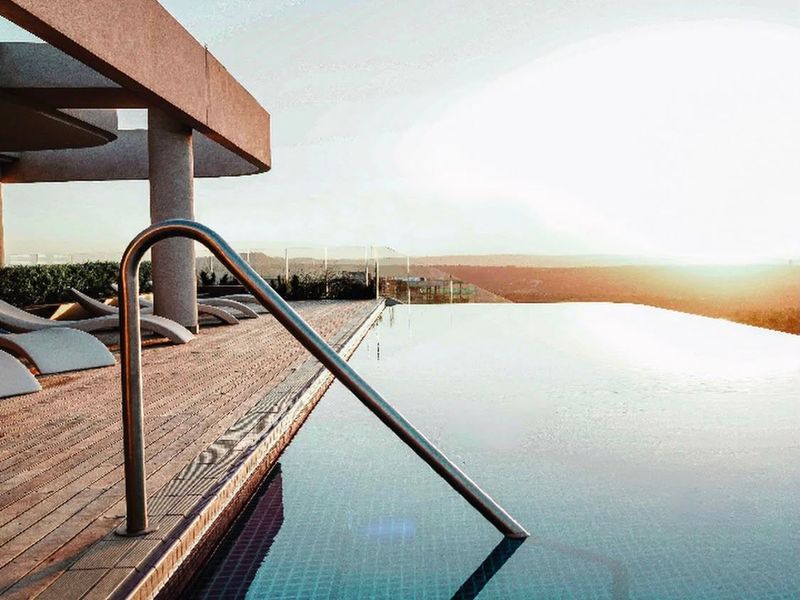An elegant infinity edge swimming pool featuring timber decking, a modern concrete pergola, and panoramic sunset views over the surrounding landscape.