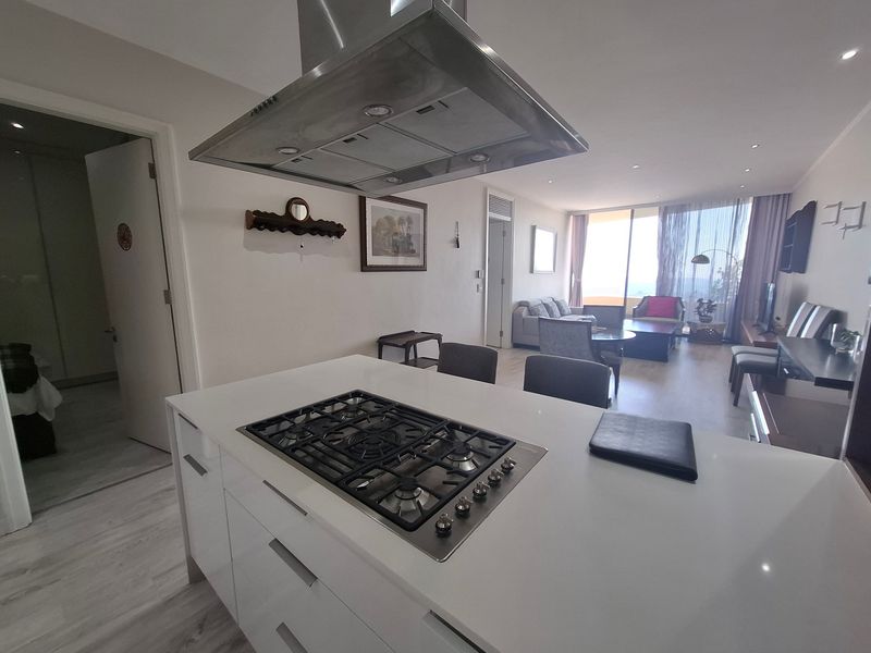 Modern open-plan living area featuring a central kitchen island with gas hob, stainless steel extractor, and seamless flow to the lounge and balcony.