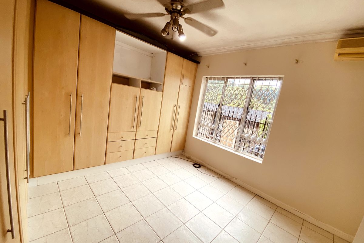 A spacious, empty bedroom featuring built-in wooden cupboards, tiled flooring, a ceiling fan, and an air conditioning unit with security bars on the window.