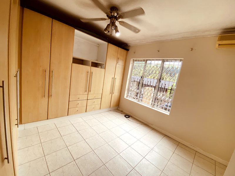 A spacious, empty bedroom featuring built-in wooden cupboards, tiled flooring, a ceiling fan, and an air conditioning unit with security bars on the window.