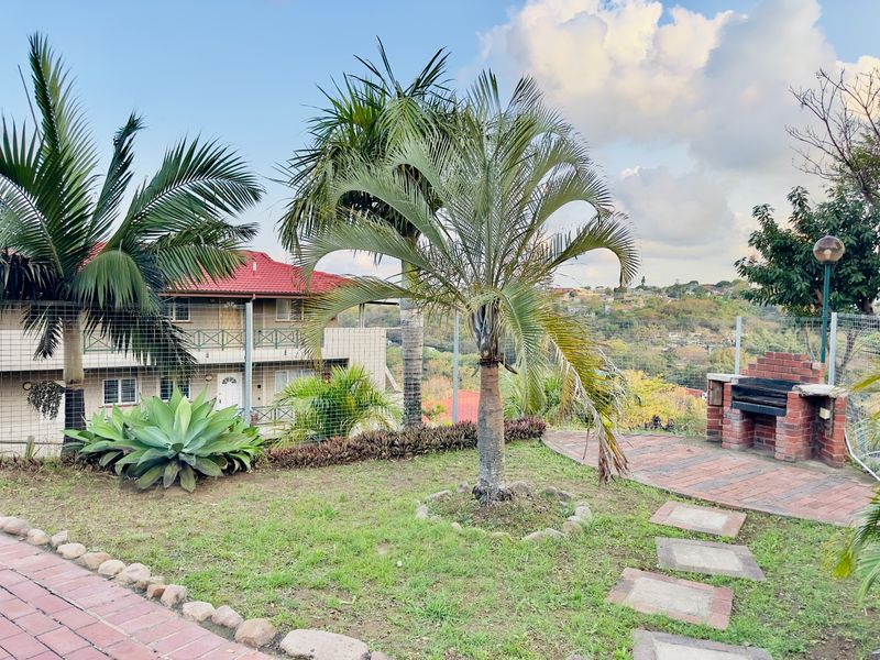 Rear garden area featuring a brick built-in braai, paved pathways, and established landscaping with views of the surrounding neighborhood.