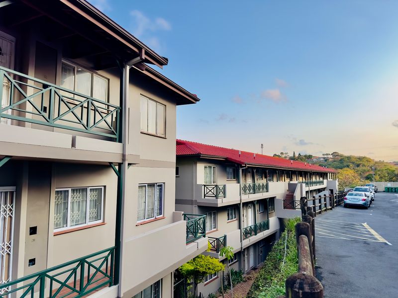 Exterior view of a multi-story apartment complex featuring balconies with green railings, tiled roofing, and an adjacent paved parking area.
