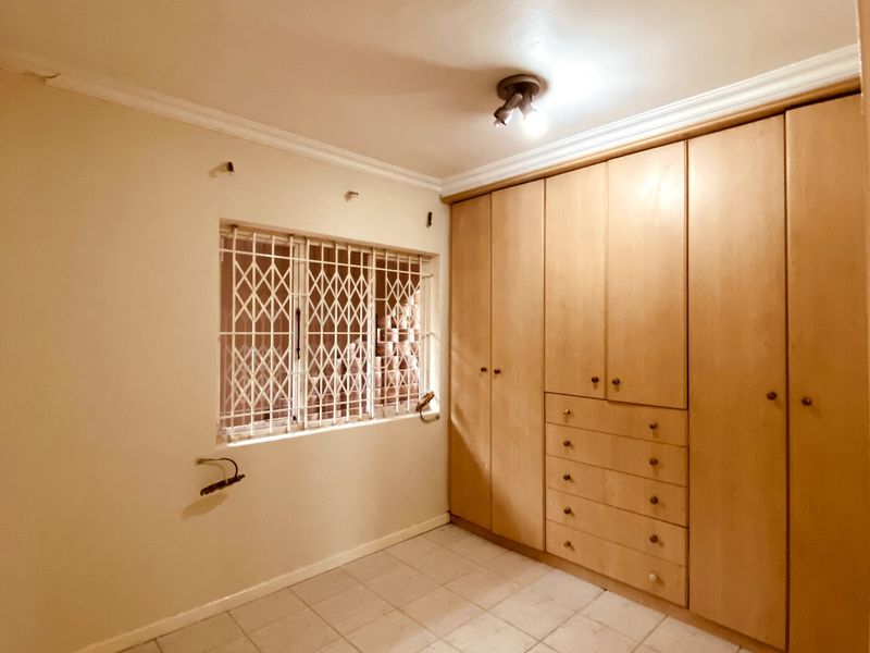 A vacant bedroom featuring built-in wooden cupboards, tiled flooring, and a window fitted with a security gate.