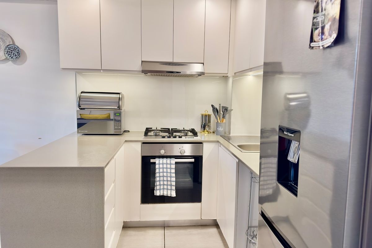 A modern, compact kitchen featuring white cabinetry, stone countertops, a gas hob, built-in oven, and a sleek glass splashback.
