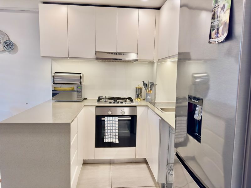 A modern, compact kitchen featuring white cabinetry, stone countertops, a gas hob, built-in oven, and a sleek glass splashback.