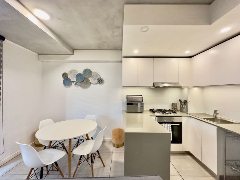 Modern open-plan kitchen and dining area featuring sleek white cabinetry, stone countertops, integrated appliances, and recessed lighting with neutral tiled flooring.