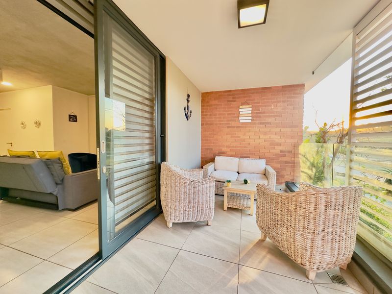 A covered balcony featuring tiled flooring, an exposed brick feature wall, and a glass balustrade, accessible via large sliding glass doors.