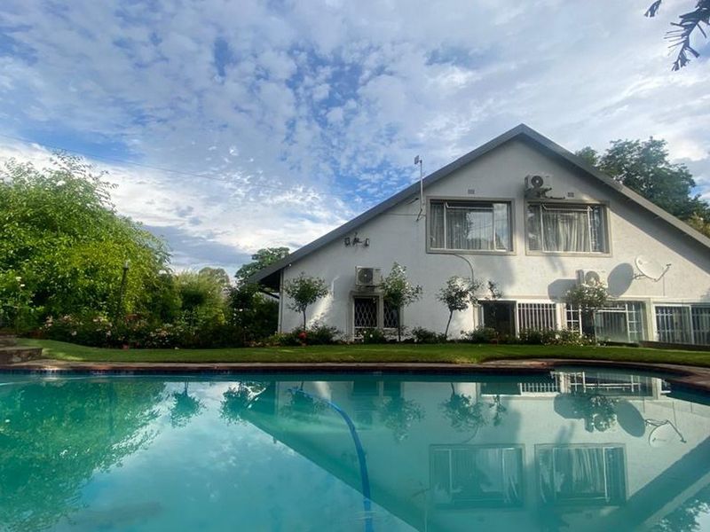 A well-maintained swimming pool area featuring a clear pool, landscaped garden, and the rear elevation of the house with mounted air conditioning units.