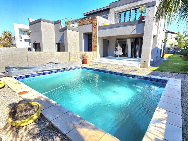 Rear exterior view featuring a swimming pool with tiled coping, stone-clad architectural accents, and a paved patio area leading to the main residence.