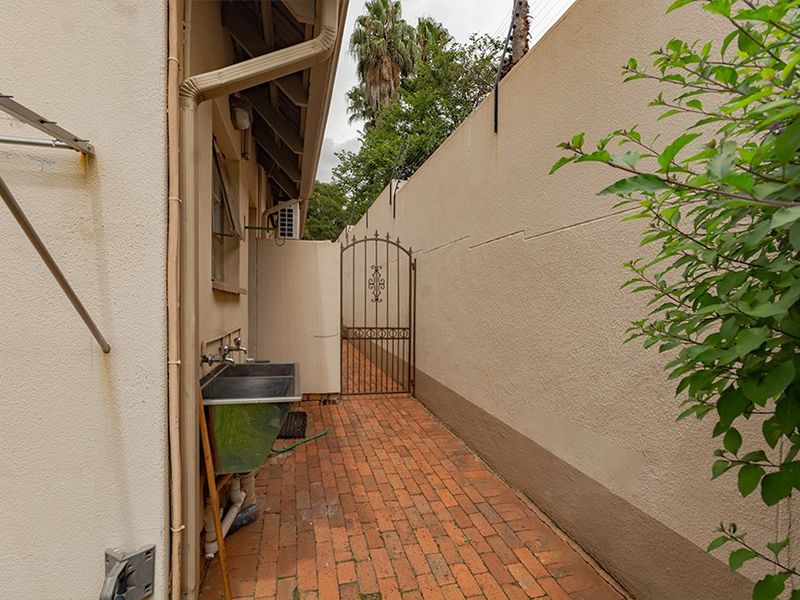 A narrow rear exterior side passage featuring brick paving, a utility sink, and a secure metal gate set against a perimeter wall.