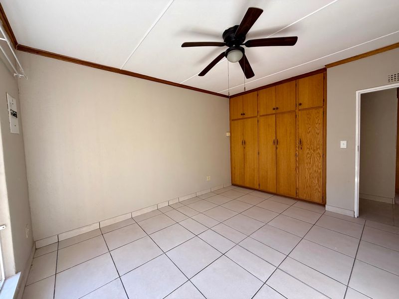 A spacious, empty bedroom featuring neutral tiled flooring, a ceiling fan, and ample built-in wooden cabinetry for storage.