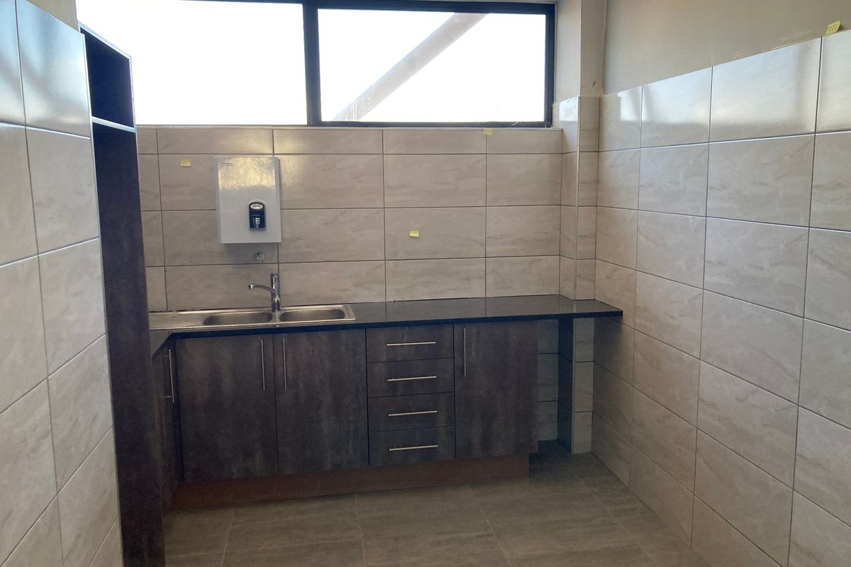 A clean, functional kitchenette area featuring tiled walls, dark wood-finish cabinetry, a stainless steel sink, and a high-level window for natural light.