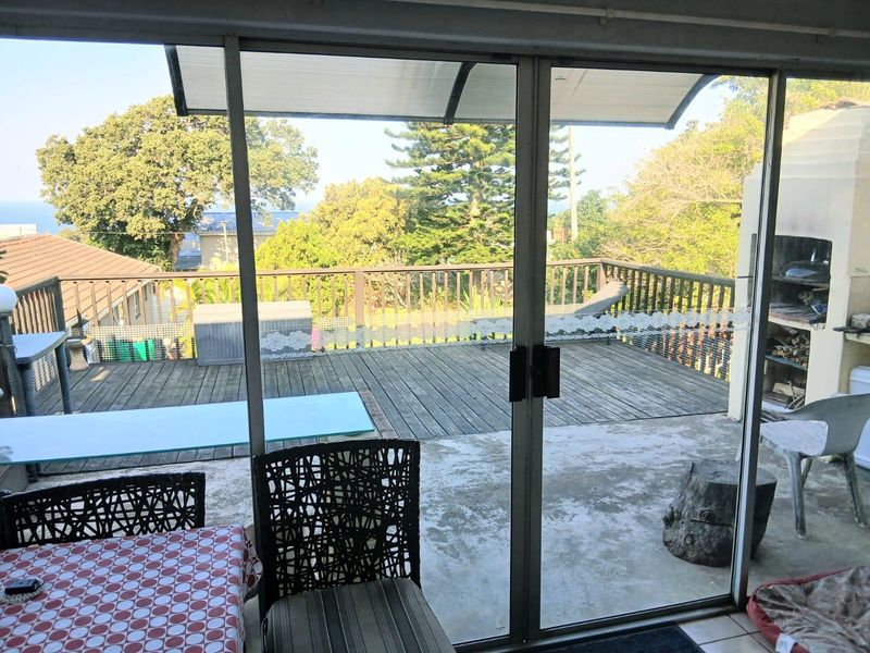 An indoor-outdoor living space featuring sliding glass doors opening onto a wooden deck with a built-in braai and garden views.