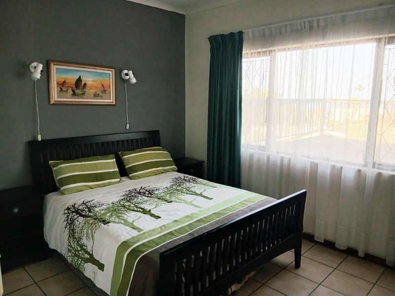 A functional bedroom featuring tiled flooring, a large window with security bars, and wall-mounted lighting, offering a comfortable and practical living space.