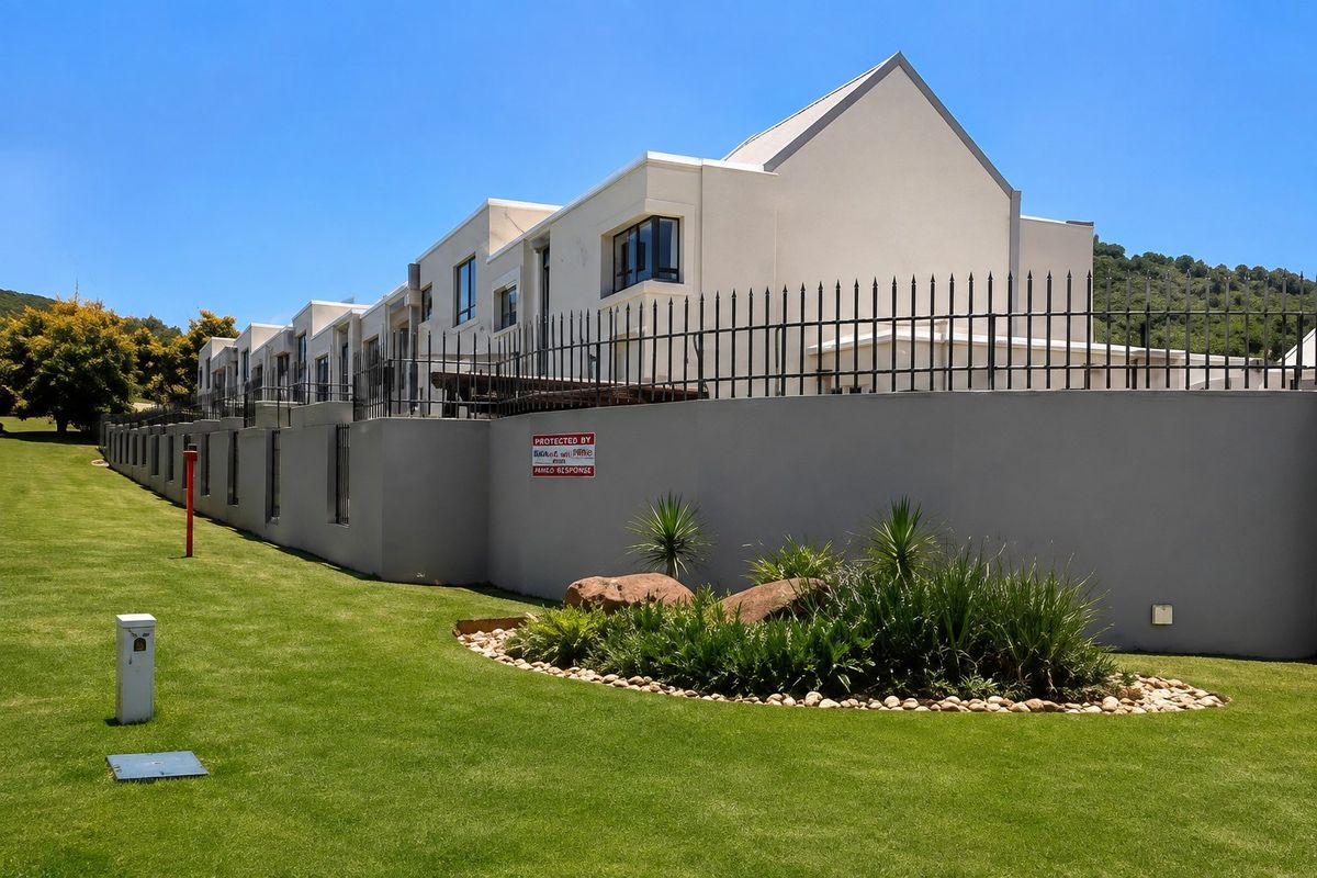 A view of the rear exterior of the townhouse complex, featuring a well-maintained lawn, landscaped garden beds, and secure perimeter wall with metal fencing.