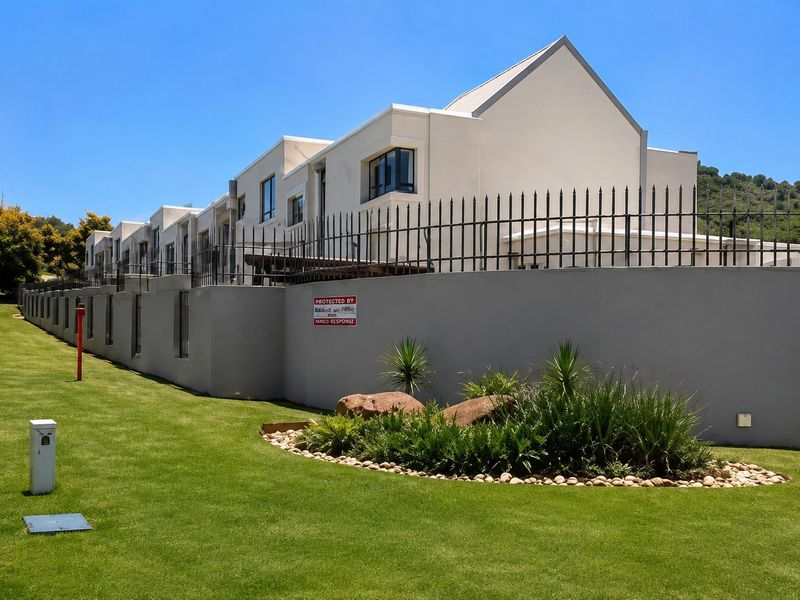 A view of the rear exterior of the townhouse complex, featuring a well-maintained lawn, landscaped garden beds, and secure perimeter wall with metal fencing.