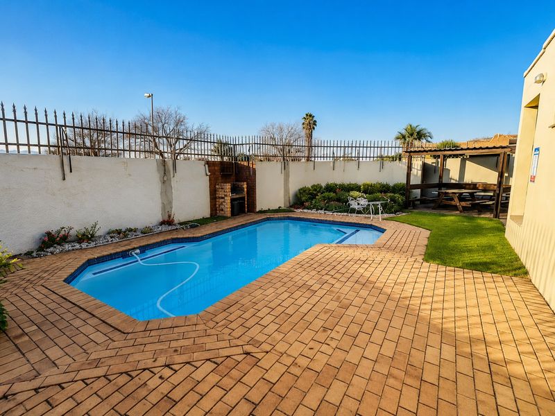 Outdoor pool area featuring a swimming pool, brick paving, built-in braai, and a pergola, enclosed by a perimeter wall with security fencing.