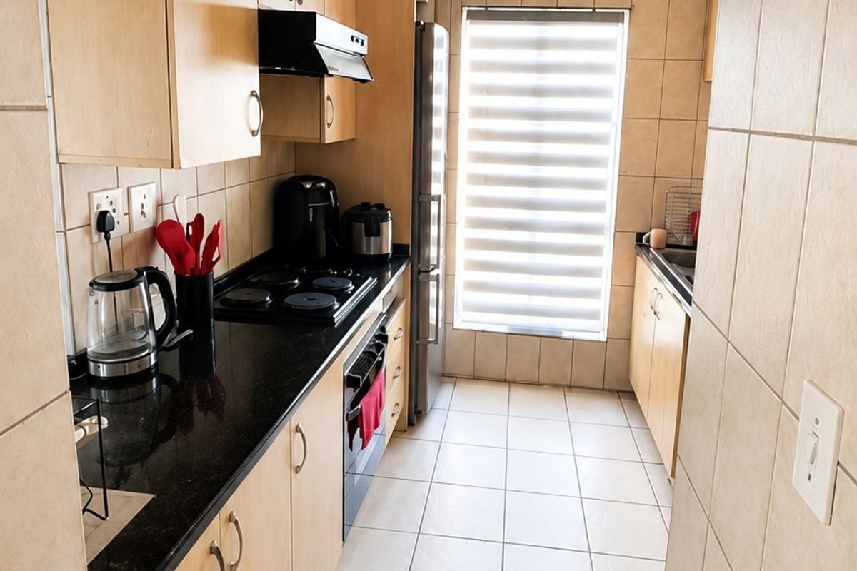 A functional galley-style kitchen featuring light wood cabinetry, dark granite countertops, tiled finishes, and integrated cooking appliances.