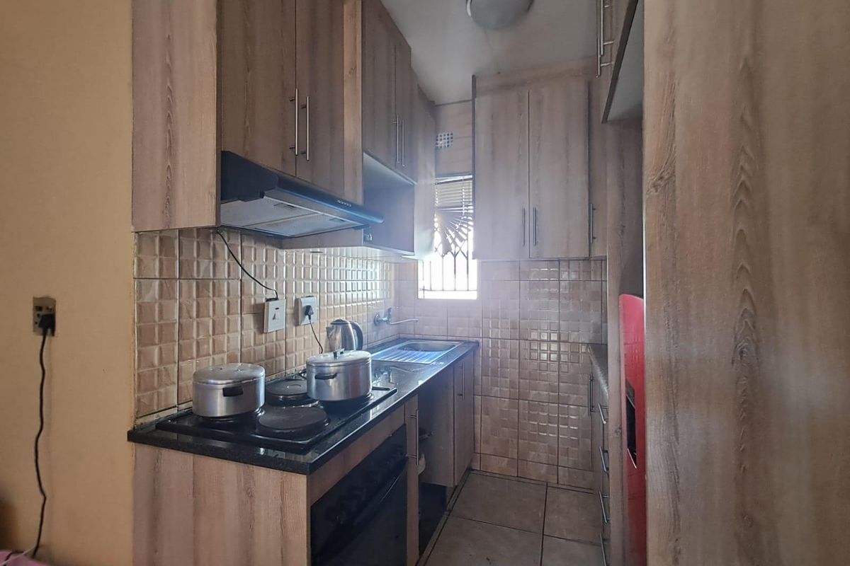 Compact kitchen featuring wood-look cabinetry, tiled backsplash, built-in oven and hob, and functional stainless steel sink with security bars on the window.