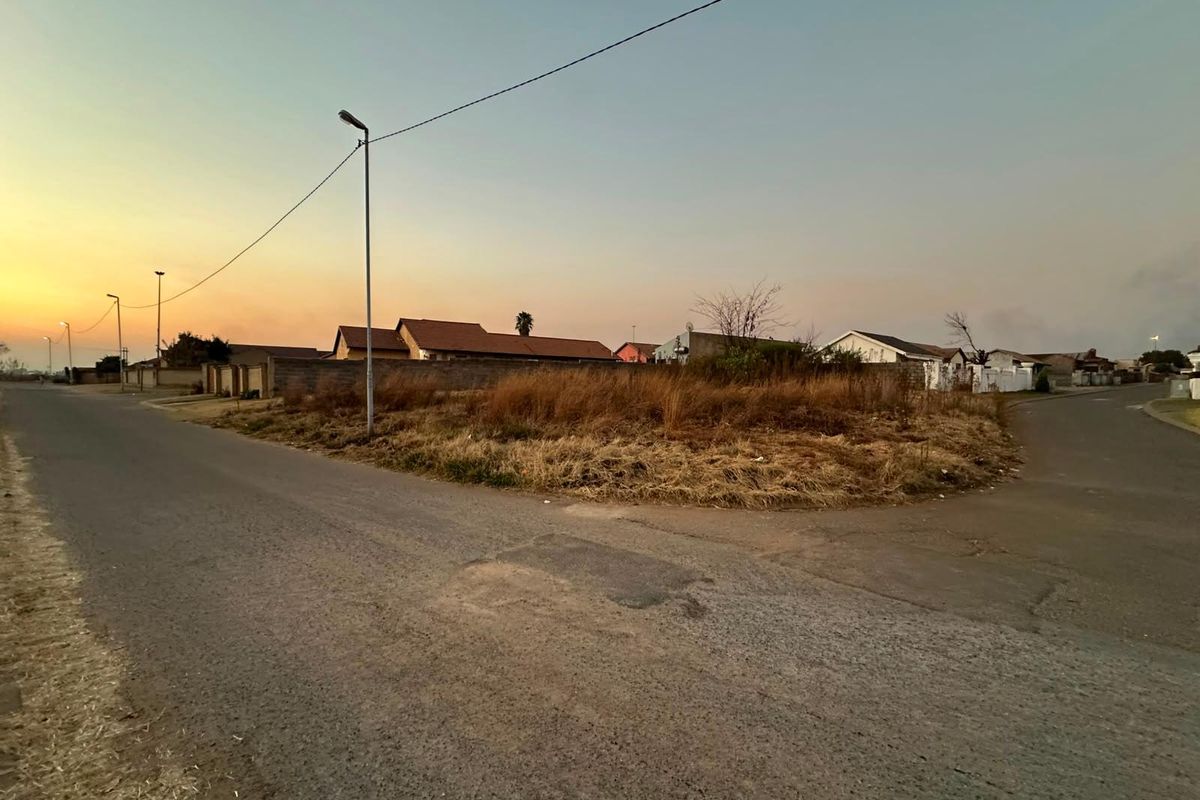 A vacant residential corner plot featuring street access, boundary walling, and established street lighting in a suburban neighborhood.