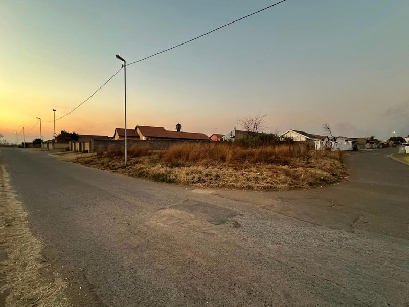 A vacant residential corner plot featuring street access, boundary walling, and established street lighting in a suburban neighborhood.