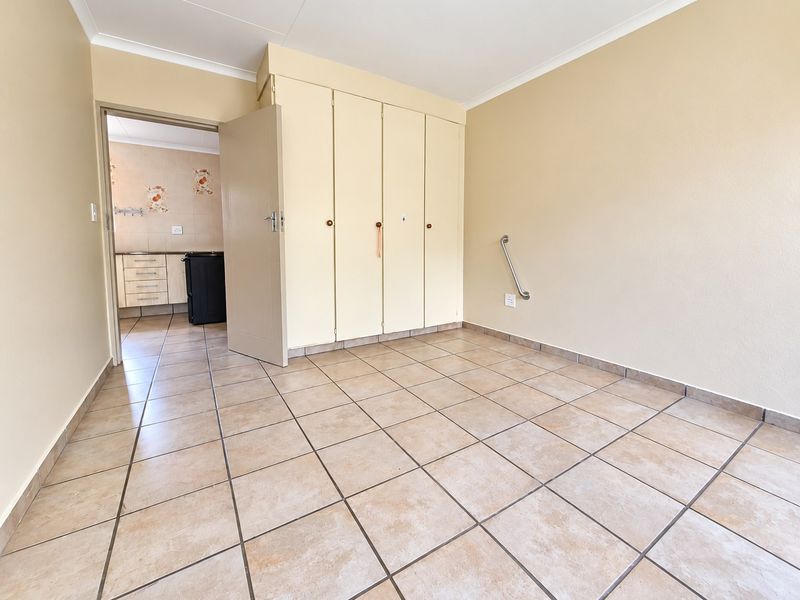 A clean, empty bedroom featuring neutral ceramic floor tiles, built-in cupboards, and a wall-mounted grab rail, with an adjacent view into the kitchen area.