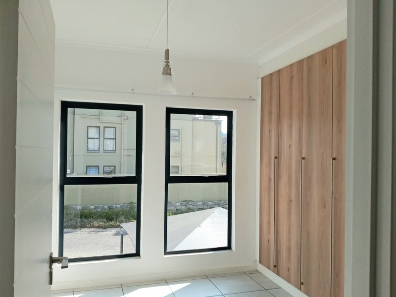 A bright, modern bedroom featuring neutral tiled flooring, built-in wooden cupboards, and large aluminium-framed windows providing ample natural light.